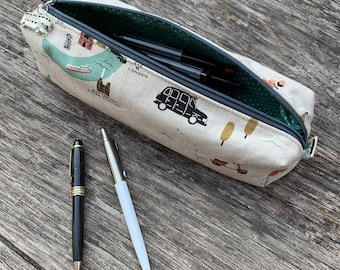 Op de afbeelding: Een beige etui met een Londen-thema print, open om pennen en potloden te laten zien. De etui heeft een ritssluiting en een groene polka dot binnenkant. Twee pennen, een zwarte en gouden, de andere zilver en wit, liggen naast de etui.