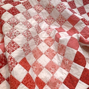 May include: A handmade quilt with a patchwork design. The quilt features a repeating pattern of red and white squares. Some red squares have floral or paisley patterns. The quilt is folded, showing the texture of the fabric.