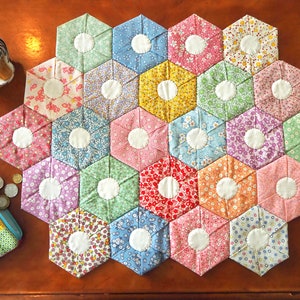 Peut inclure: Une courtepointe patchwork faite de pièces de tissu hexagonales aux couleurs pastel variées. Chaque hexagone présente un motif floral et un centre circulaire blanc. Un pot à crayons et une trousse sont également visibles.