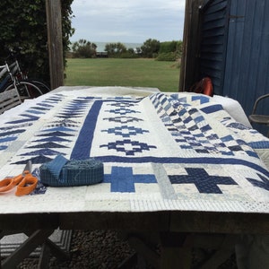 Peut inclure: Une courtepointe blanche avec des carrés et des triangles à motifs bleus et blancs est posée sur une table en bois. La courtepointe est en cours de préparation pour la reliure. Une paire de ciseaux et un rouleau de tissu bleu sont sur la table.