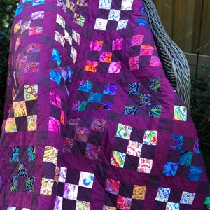 Może przedstawiać: Ręcznie robiona kołdra z głębokim fioletowym tłem i wzorem patchworku. Kołdra posiada kwadraty z różnych kolorowych tkanin kwiatowych i wzorzystych, tworząc żywy i teksturowany wygląd. Kołdra jest narzucona na wiklinowy fotel.