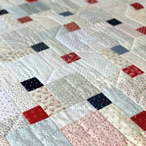 Peut inclure: Une courtepointe en patchwork avec une variété de tissus à motifs rouges, blancs et bleus. La courtepointe est composée de carrés et de rectangles, et les coutures sont visibles. La courtepointe est matelassée avec un motif de points simple.