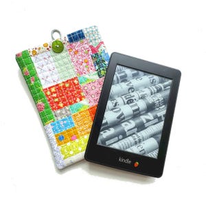 Op de afbeelding: Een zwarte Kindle e-reader met krantentekst op het scherm ligt naast een kleurrijke patchwork stoffen hoes. De hoes heeft verschillende patronen in rood, groen, blauw en roze, met een groene knoop en een zilveren lus.