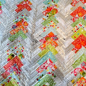 May include: A colorful quilt with a herringbone pattern. The quilt features a variety of fabrics in shades of red, orange, green, blue, and gray. Some fabrics have a print of text. The quilt is machine-stitched with white thread.