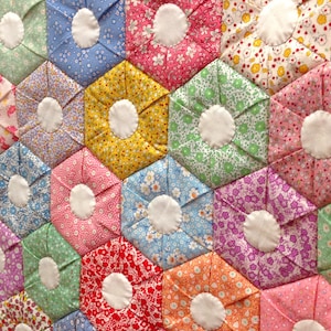 May include: Close-up of a handmade quilt with a floral pattern. The quilt is composed of hexagonal fabric pieces in various colors, including blue, pink, yellow, and green, each with a white circular center. The quilt's design is reminiscent of a flower.
