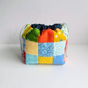Pode incluir: Uma colorida bolsa com cordão de patchwork. A bolsa é feita de quadrados de tecido em tons de azul, amarelo, laranja, verde e vermelho. A parte superior da bolsa é de tecido jeans azul escuro e tem um cordão verde claro.