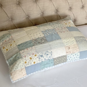 Könnte beinhalten: Ein rechteckiger Patchwork-Kissenbezug mit verschiedenen Stoffmustern in Blau-, Creme- und Grautönen. Der Kissenbezug hat ein gestepptes Design mit kleinen Quadraten und einem gemusterten Rand. Das Kissen ist ca. 70 cm lang.