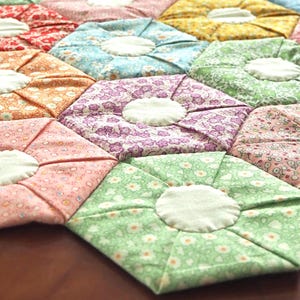 May include: Close-up of a handmade quilt with a floral pattern. The quilt features hexagonal shapes in various colors, including red, orange, pink, blue, green, and purple. Each hexagon has a white circular center.