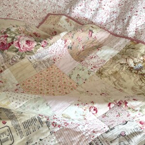 K&ouml;nnte beinhalten: Ein Patchwork-Quilt in Rosa-, Creme- und Beiget&ouml;nen mit floralen und Schriftmustern. Der Quilt zeigt Rosen, Streifen und franz&ouml;sischen Text, was ihm einen Vintage-Look verleiht. Die R&auml;nder sind mit einem rosa Saum versehen.
