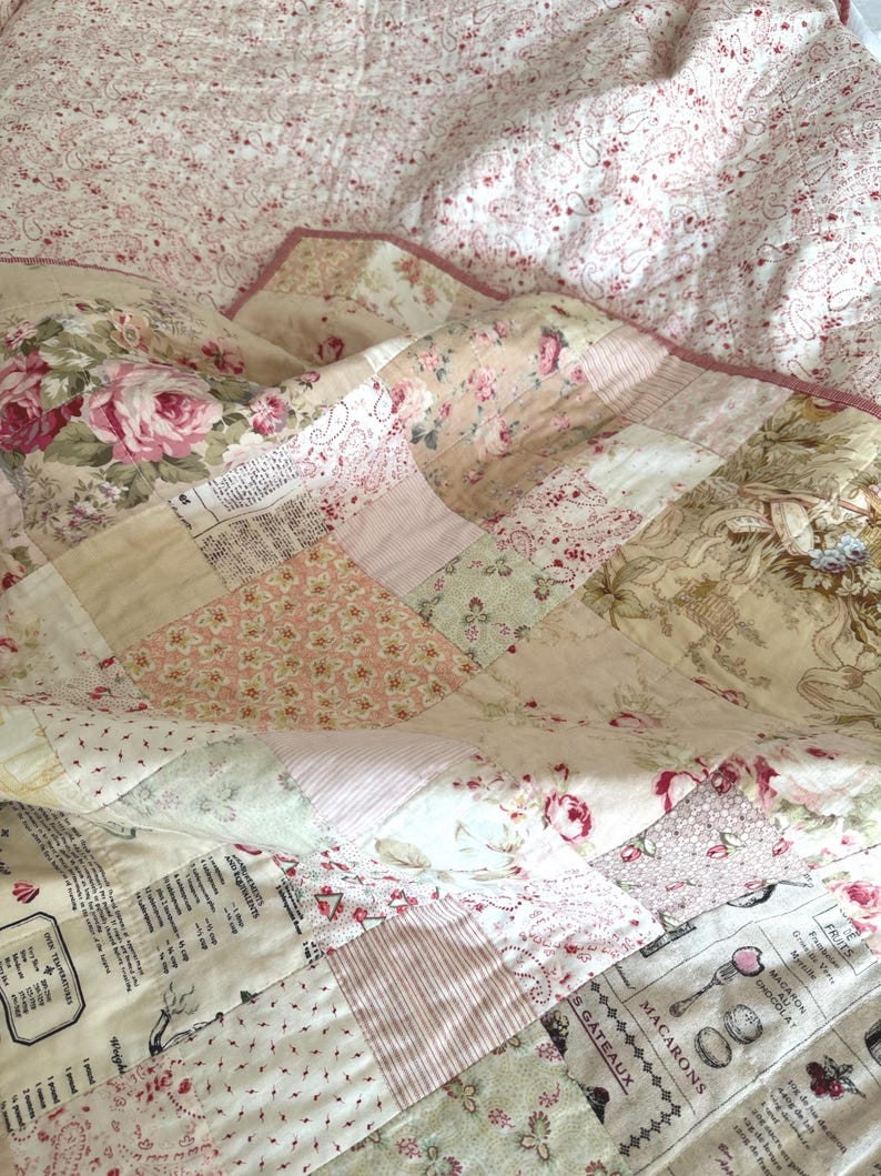 K&ouml;nnte beinhalten: Ein Patchwork-Quilt in Rosa-, Creme- und Beiget&ouml;nen mit floralen und Schriftmustern. Der Quilt zeigt Rosen, Streifen und franz&ouml;sischen Text, was ihm einen Vintage-Look verleiht. Die R&auml;nder sind mit einem rosa Saum versehen.