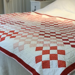 Op de afbeelding: Een handgemaakte quilt met een dambordpatroon in tinten rood en wit. De quilt heeft een rode rand met een wit en rood gestreepte bies. De quilt ligt op een bed met een witte bedrok.