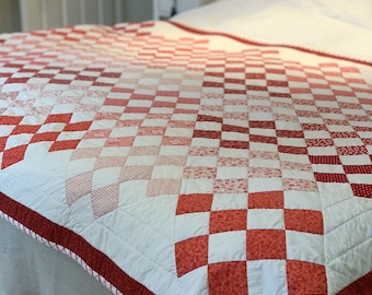 May include: A handmade quilt with a checkerboard pattern in shades of red and white. The quilt features a red border with a white and red striped trim. The quilt is placed on a bed with a white bed skirt.