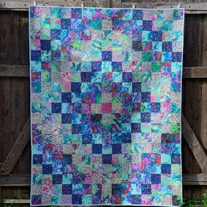 Op de afbeelding: Een kleurrijk quilt met een patchwork-ontwerp met vierkanten in verschillende tinten blauw, groen, roze en paars. De quilt hangt aan een houten hek.