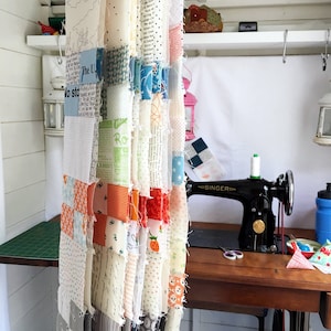 May include: A close-up of a fabric quilt being sewn together. The quilt is made of many different colorful fabrics, including white, pink, orange, and blue. The quilt is hanging from a hook in a room with a sewing machine and other sewing supplies.