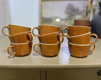 Pode incluir: Seis chávenas de café de cerâmica castanha empilhadas em pares, com acabamento branco. Cada chávena tem uma pega arredondada. As chávenas estão dispostas em três pilhas sobre uma superfície clara. O fundo inclui um quadro emoldurado e objetos decorativos.