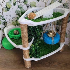 May include: A multi-tiered white and rope hermit crab habitat with artificial grass, a ramp, and a blue tire swing. The habitat includes a green turtle-shaped dish and several hermit crab shells. The structure is decorated with faux greenery.