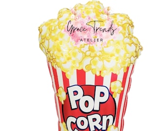 Può includere: Un palloncino a forma di popcorn con un contenitore a righe rosse e bianche. Il palloncino è pieno di popcorn gialli e presenta le parole "POP CORN" in un cerchio rosso. È visibile anche il testo "Grace Trends ATELIER".