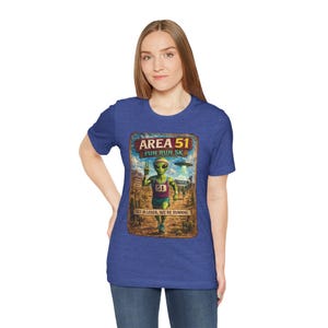 Puede incluir: Una camiseta azul de manga corta con un estampado gráfico de un alienígena verde corriendo en un paisaje desértico. El texto dice Area 51 Fun Run 5K y Get in Loser, We're Running. El alienígena lleva una camiseta sin mangas morada con el número 51.