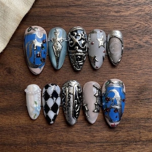 May include: A collection of ten artificial nails with various designs. The nails feature a range of colors, including blue, silver, black, and white. Designs include floral patterns, geometric shapes, and metallic accents. The nails are oval-shaped.