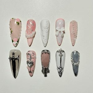 May include: A collection of ten long, almond-shaped artificial nails. Designs include pink and white with floral, bow, and pearl accents, and silver and black with cross and heart details. Some nails have a glittery finish.