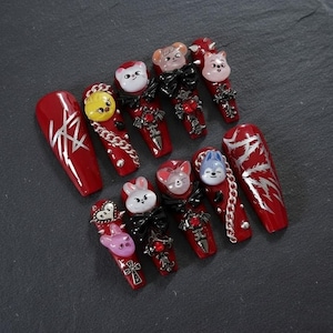 May include: A set of red coffin-shaped press-on nails. Some nails feature silver designs, while others have 3D animal faces, black bows, and silver chain accents. The nails are decorated with small red gems and crosses.