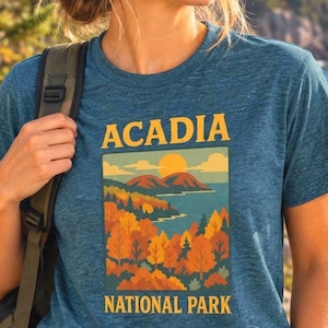 May include: A heathered blue short-sleeve t-shirt featuring a graphic print of Acadia National Park. The design displays yellow text, a bright sun, mountains, and a forest with orange and green trees above the words National Park.
