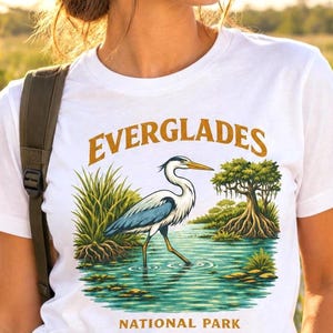 May include: A white short-sleeved t-shirt featuring a graphic print of a blue and white heron standing in water with green marsh grass and a cypress tree. The text EVERGLADES is printed in gold above the bird and NATIONAL PARK below it.