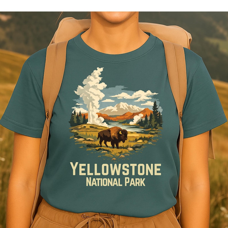 May include: A teal short-sleeved t-shirt featuring a graphic illustration of a bison standing in a landscape with a geyser, mountains, and trees. The text "YELLOWSTONE NATIONAL PARK" is printed in a light cream color below the graphic design.