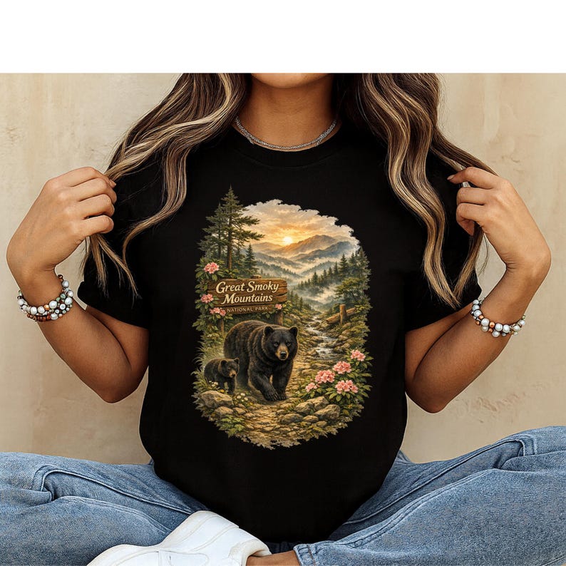 May include: A black t-shirt featuring a graphic print of a mother bear and cub walking on a path in the Great Smoky Mountains National Park. The scene includes a wooden sign, pine trees, a flowing stream, and pink flowers under a sunset sky.