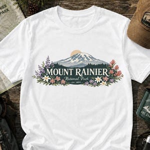 May include: A white short-sleeved t-shirt featuring a graphic of Mount Rainier with a sun, forest, and colorful wildflowers. The text reads MOUNT RAINIER National Park Est. 1899. The design is centered on the chest of the shirt.