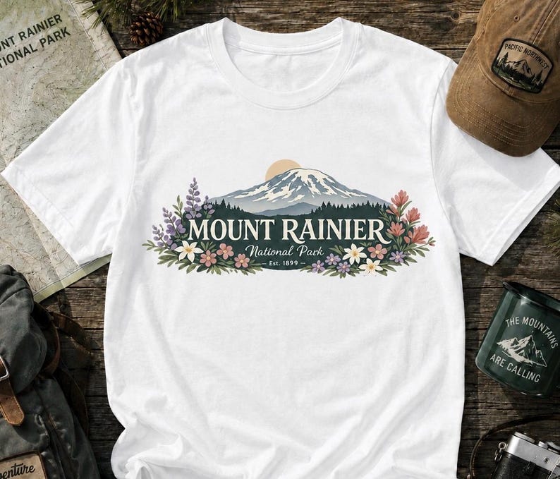 May include: A white short-sleeved t-shirt featuring a graphic of Mount Rainier with a sun, forest, and colorful wildflowers. The text reads MOUNT RAINIER National Park Est. 1899. The design is centered on the chest of the shirt.