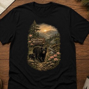 May include: A black short-sleeve t-shirt featuring a graphic print of a black bear and a cub walking on a rocky path in the Great Smoky Mountains National Park, with a sunset, pine trees, and pink flowers surrounding the scene.