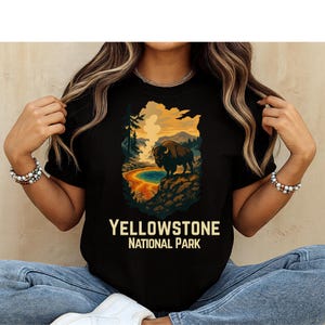 May include: A black t-shirt featuring a graphic illustration of a bison standing on a rocky ledge overlooking a geothermal pool and mountains. The text "YELLOWSTONE NATIONAL PARK" is printed in a bold, light-colored font below the graphic.