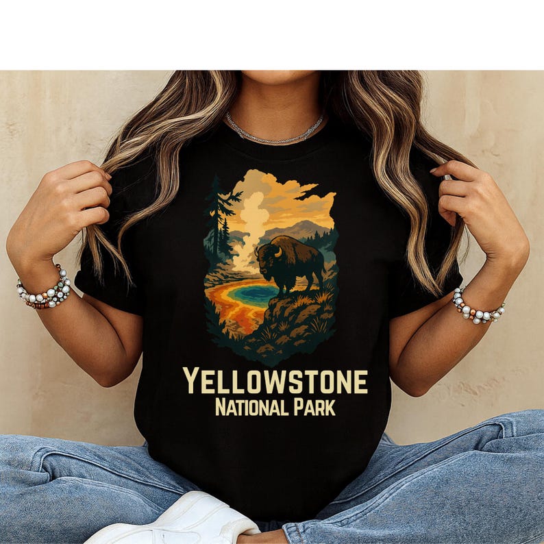 May include: A black t-shirt featuring a graphic illustration of a bison standing on a rocky ledge overlooking a geothermal pool and mountains. The text "YELLOWSTONE NATIONAL PARK" is printed in a bold, light-colored font below the graphic.
