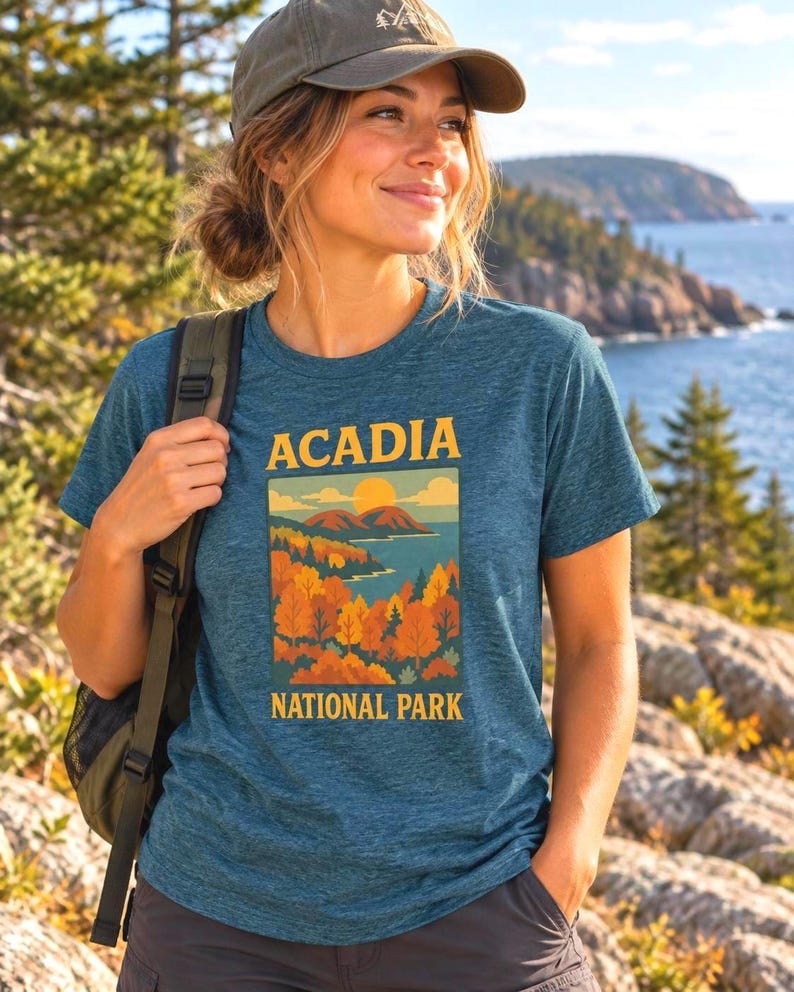 May include: A heathered blue short-sleeve t-shirt featuring a graphic print of Acadia National Park. The design displays yellow text, a bright sun, mountains, and a forest with orange and green trees above the words National Park.