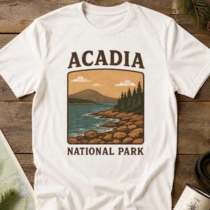 May include: A white short-sleeve t-shirt featuring a graphic print of a coastal landscape with mountains, pine trees, and a rocky shoreline. The text ACADIA is printed above the image and NATIONAL PARK is printed below in dark brown lettering.