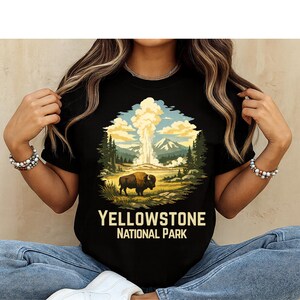 May include: A black t-shirt featuring a graphic illustration of a bison standing in a landscape with a geyser erupting in the background, surrounded by pine trees and mountains. The text "YELLOWSTONE NATIONAL PARK" is printed in yellow below the image.
