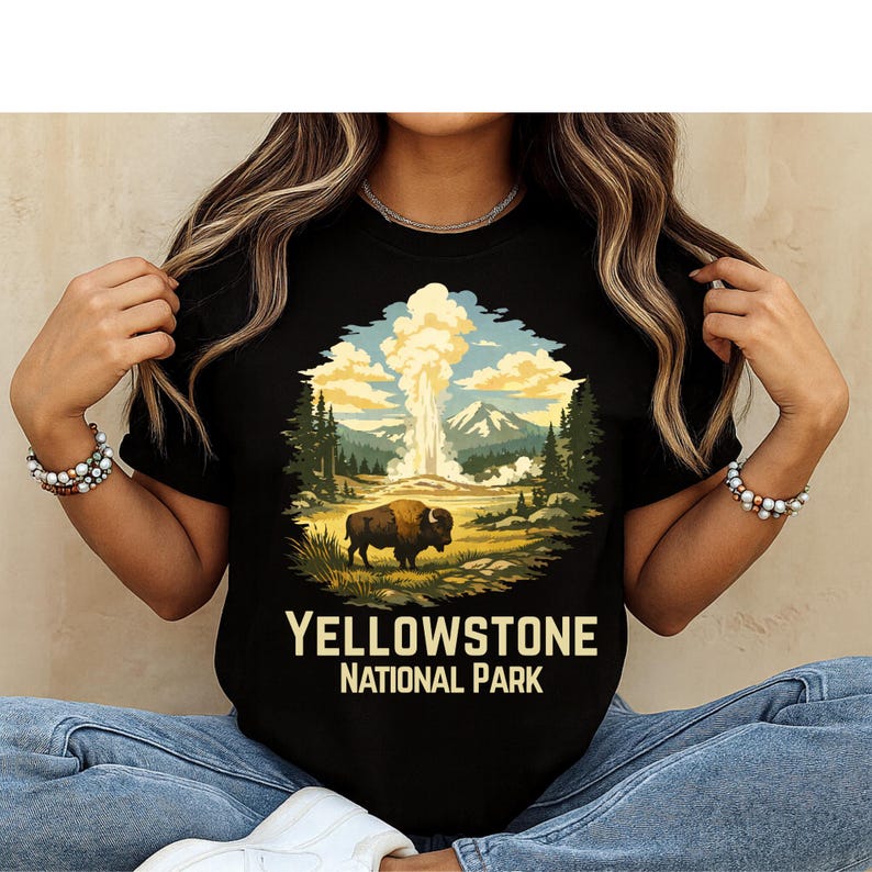 May include: A black t-shirt featuring a graphic illustration of a bison standing in a landscape with a geyser erupting in the background, surrounded by pine trees and mountains. The text "YELLOWSTONE NATIONAL PARK" is printed in yellow below the image.