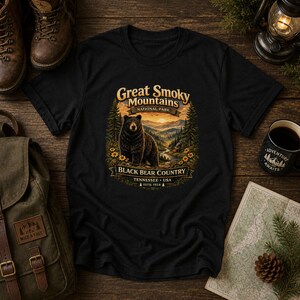 May include: A black short-sleeve t-shirt featuring a graphic print of a black bear in a mountain landscape. The text reads Great Smoky Mountains National Park, Black Bear Country, Tennessee, USA, ESTD. 1934, surrounded by pine trees and yellow flowers.