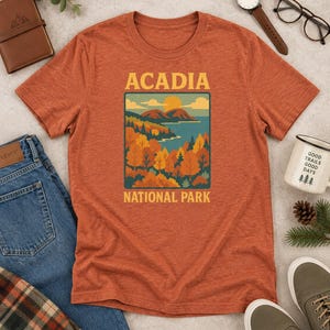 May include: A heathered rust-orange short-sleeve t-shirt featuring a graphic print of a landscape with mountains, a lake, and autumn trees. The text ACADIA is printed above the graphic, and NATIONAL PARK is printed below in a yellow sans-serif font.