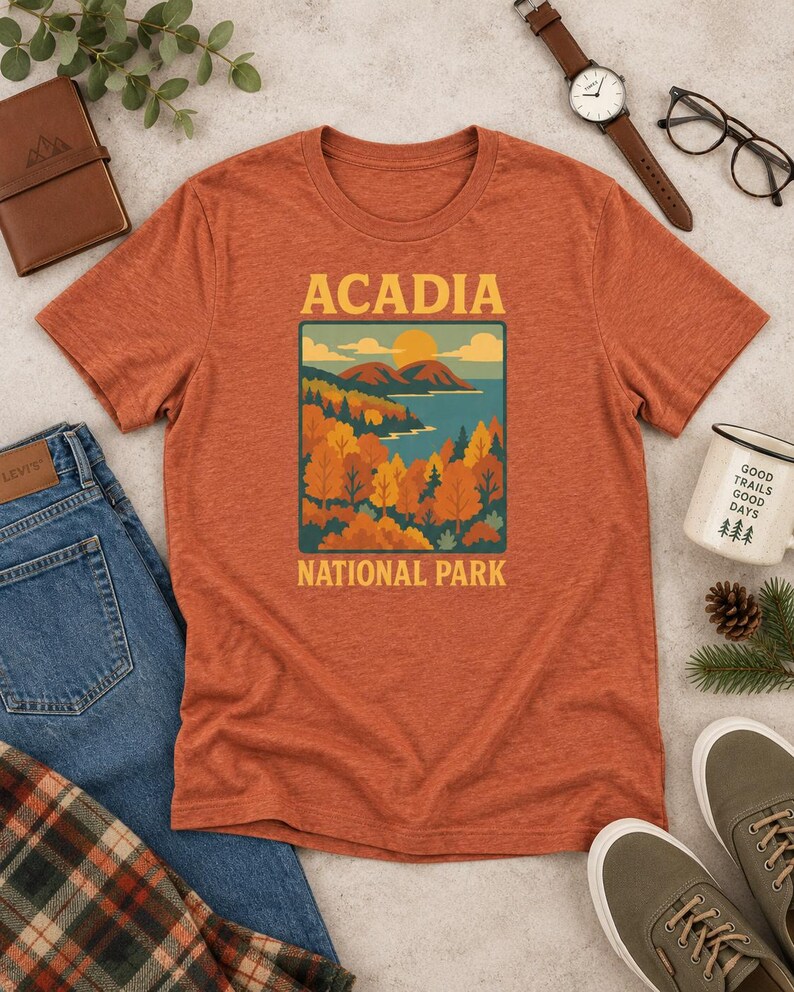 May include: A heathered rust-orange short-sleeve t-shirt featuring a graphic print of a landscape with mountains, a lake, and autumn trees. The text ACADIA is printed above the graphic, and NATIONAL PARK is printed below in a yellow sans-serif font.