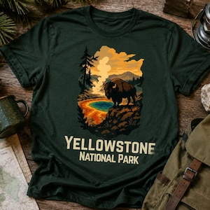 May include: A dark green short-sleeve t-shirt featuring a graphic illustration of a bison standing on a rocky cliff overlooking a colorful geothermal pool and geyser, with the text YELLOWSTONE NATIONAL PARK printed in a bold, light-colored font below.