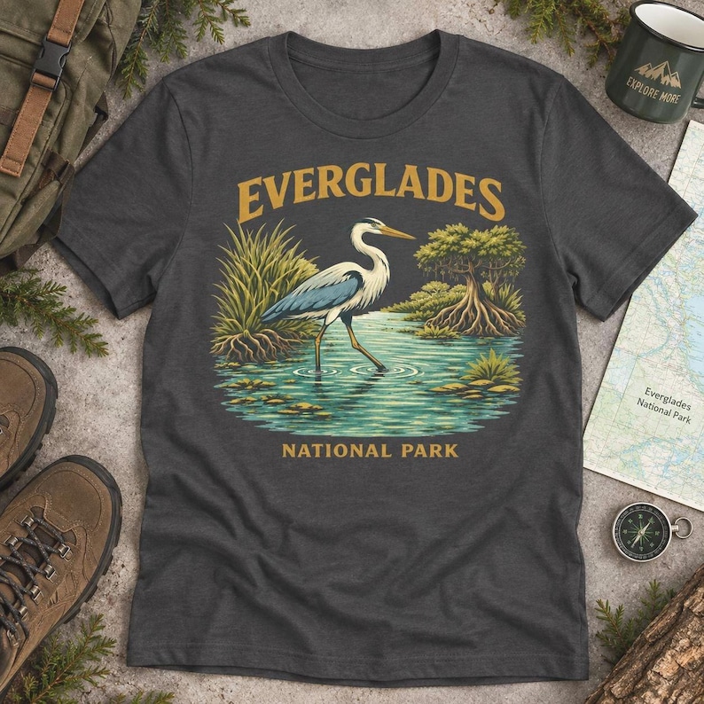 May include: A charcoal gray short-sleeve t-shirt featuring a graphic of a heron standing in water with marsh grass and a mangrove tree. The text "EVERGLADES" is printed in gold above the bird, and "NATIONAL PARK" is printed below the water.