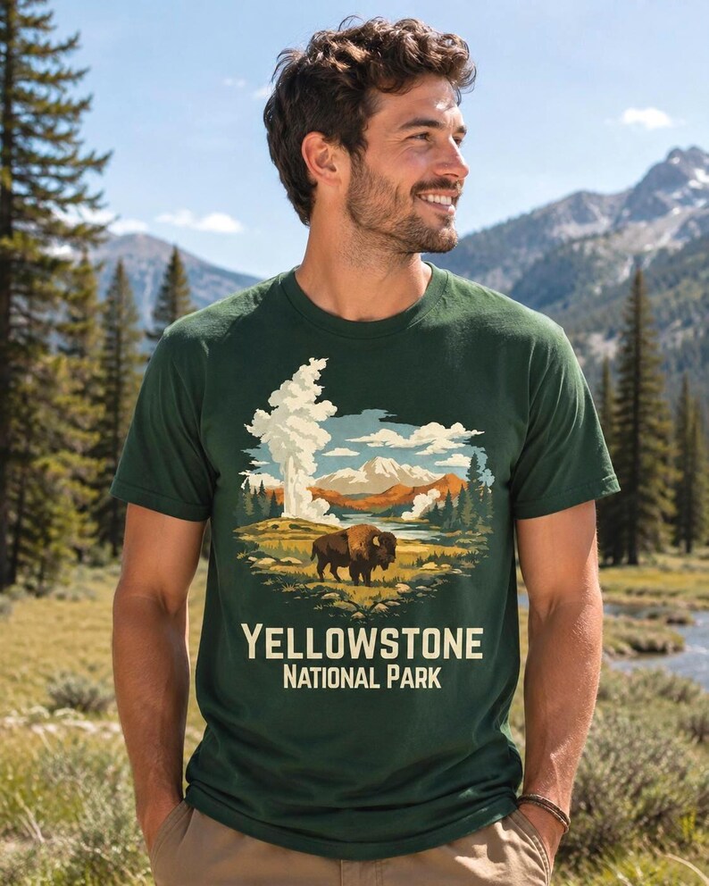 May include: A dark green short-sleeve t-shirt featuring a graphic illustration of a bison standing in a landscape with geysers, mountains, and trees. The text "YELLOWSTONE NATIONAL PARK" is printed in a cream-colored font below the illustration.