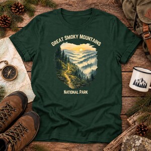 May include: A dark green t-shirt featuring a graphic of the Great Smoky Mountains National Park. The design shows a winding trail through a misty, forested mountain landscape at sunrise, with the text Great Smoky Mountains National Park.