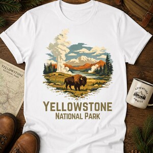 May include: A white short-sleeve t-shirt featuring a graphic illustration of a brown bison standing in a landscape with geysers, mountains, and pine trees. The text "YELLOWSTONE NATIONAL PARK" is printed in brown letters below the illustration.