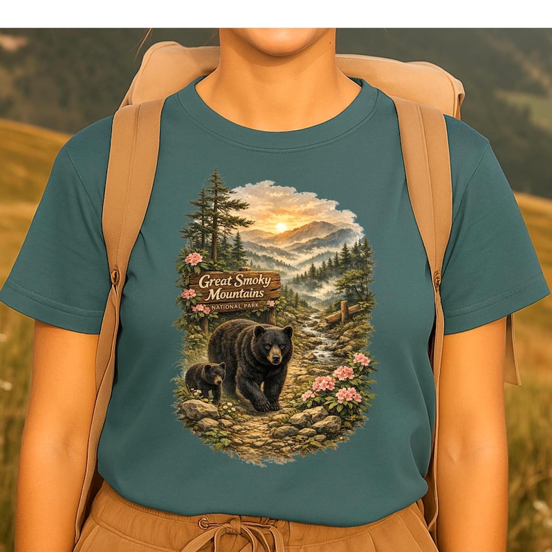 May include: A teal short-sleeve t-shirt featuring a graphic print of a black bear and cub walking on a rocky path. The scene includes a wooden sign reading Great Smoky Mountains National Park, pine trees, pink flowers, and a mountain sunset.