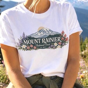 May include: A white short-sleeve t-shirt featuring a graphic of Mount Rainier with a sun, pine trees, and colorful wildflowers. The text reads MOUNT RAINIER National Park Est. 1899 in a dark font centered over the mountain illustration.