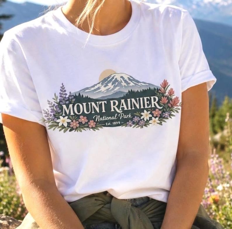 May include: A white short-sleeve t-shirt featuring a graphic of Mount Rainier with a sun, pine trees, and colorful wildflowers. The text reads MOUNT RAINIER National Park Est. 1899 in a dark font centered over the mountain illustration.