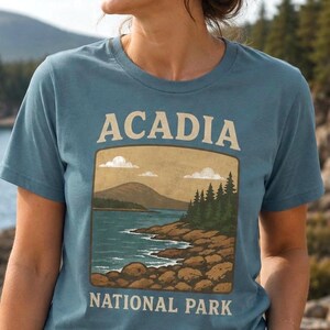 May include: A slate blue crew neck t-shirt featuring a graphic print of a coastal landscape with the text ACADIA NATIONAL PARK. The illustration depicts a rocky shoreline, a body of water, a mountain in the distance, and pine trees under a sky.